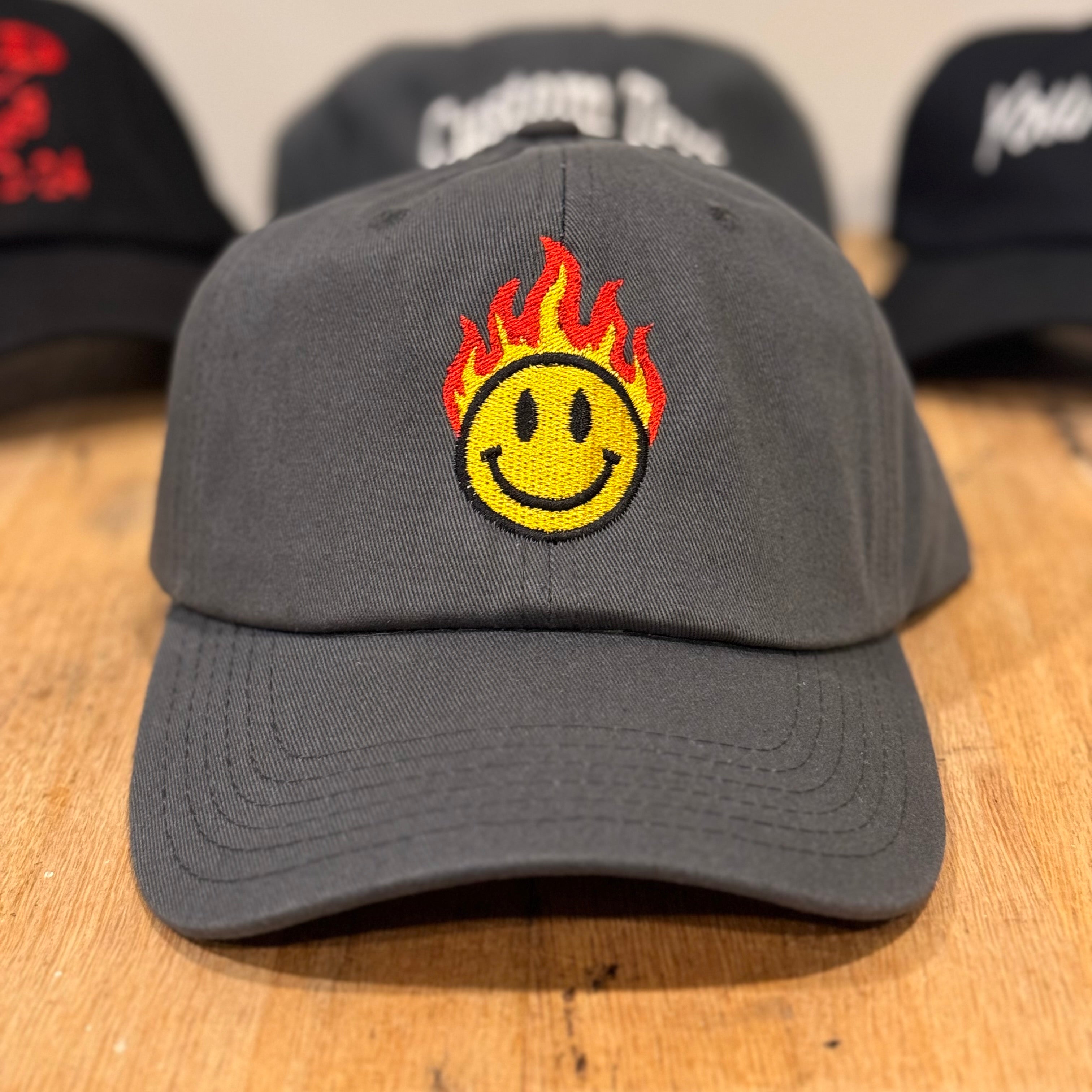 Gray cap with a smiley face design on fire, placed on a wooden surface with other caps in the background.